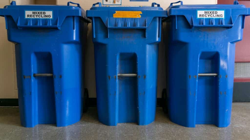 Three Large Blue Plastic Bins Lined Up Against An Indoor Wall, Each Labeled &Amp;Quot;Mixed Recycling&Amp;Quot; With Black Wheels At The Bottom And Metal Lifting Bars Across The Front.