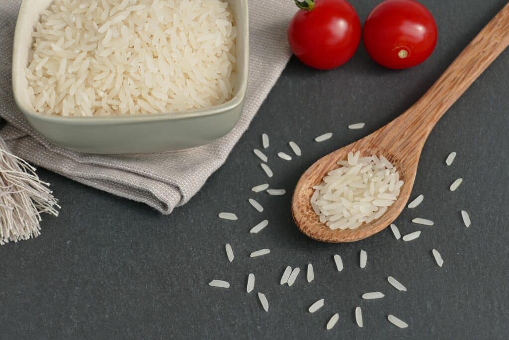 A Top-Down View Of Uncooked Basmati Rice In A Ceramic Bowl And Wooden Spoon, With Scattered Grains On A Dark Countertop. The Setup Includes A Folded Linen Cloth And Two Fresh Red Cherry Tomatoes For Contrast.