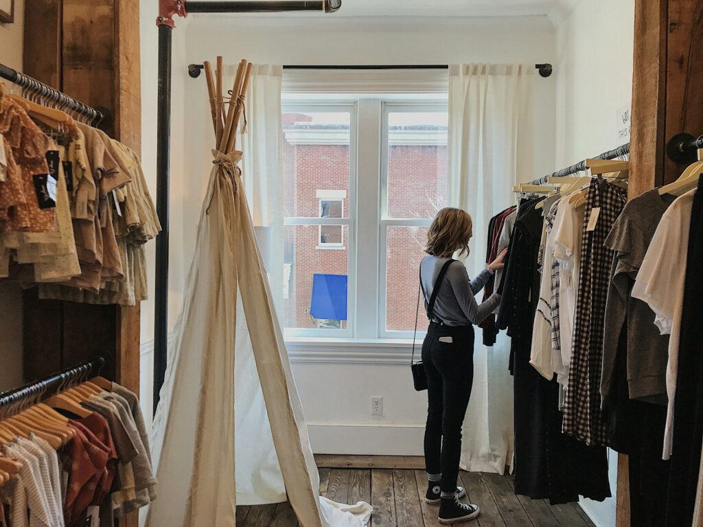 A Woman Browsing Through Clothing Racks In A Boutique Shop With Wooden Floors And A Rustic Interior. The Store Features A Mix Of Neutral-Colored And Plaid Garments On Hangers, A White Canvas Teepee, And Large Windows With White Curtains Allowing Natural Light Inside.