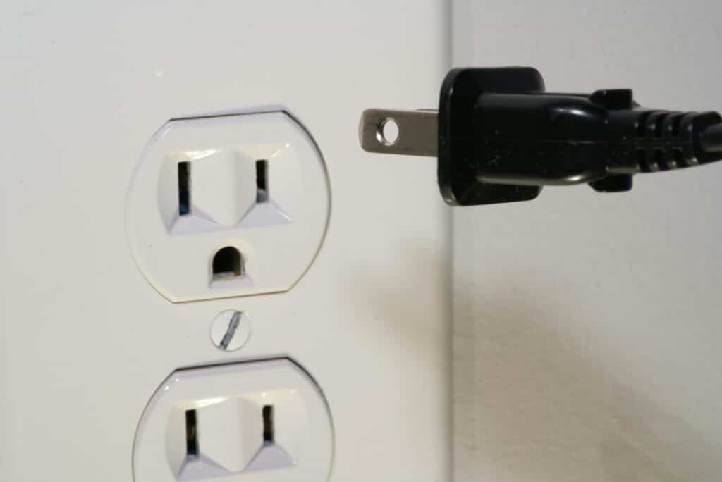 A Close-Up Image Of A Black Electrical Plug Positioned Near A White Dual Wall Outlet, Partially Inserted, With A Smooth White Wall Background.