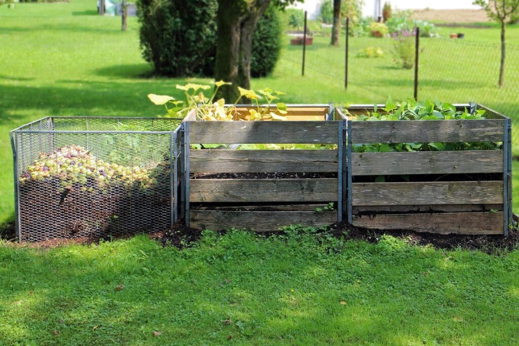 Three Outdoor Compost Bins Made Of Wooden Slats And Wire Mesh, Filled With Organic Waste And Growing Plants, Situated On A Green Lawn.