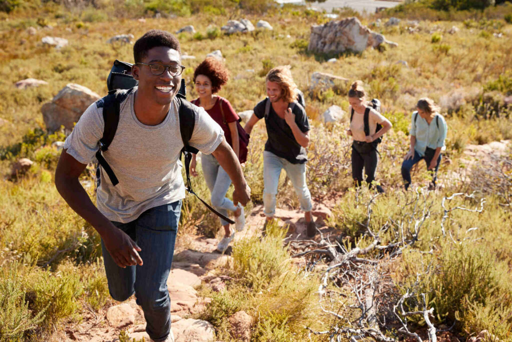 A Group Of Five People Hiking Uphill In A Sunny, Grassy Area, With Rocks And Bushes Around Them.