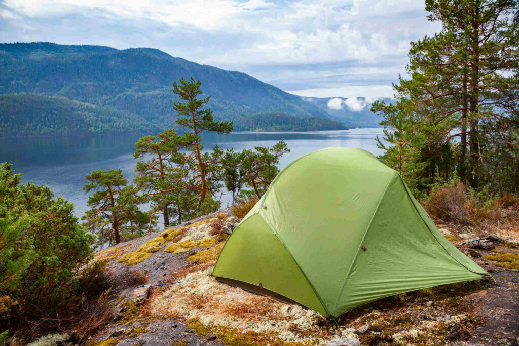 Green Tent Set Up On A Rocky Terrain Overlooking A Calm Lake Surrounded By Forested Hills Under A Cloudy Sky