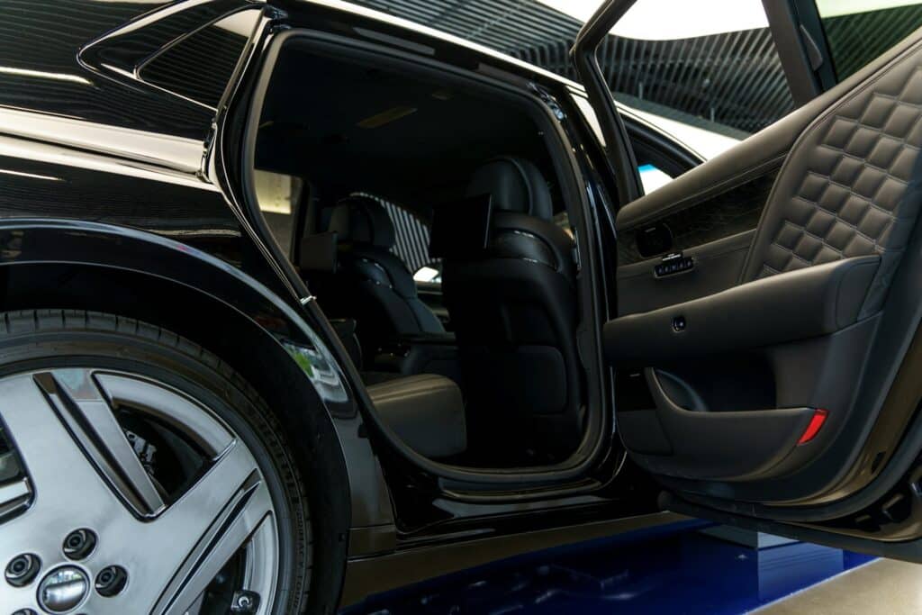 An Angled View Of An Open Luxury Car Door Revealing A High-End Black Leather Interior With Quilted Stitching, Spacious Rear Seats, And Built-In Entertainment Screens Mounted Behind The Front Seats. The Exterior Of The Vehicle Is Glossy Black With Chrome Wheel Details, Parked Indoors Under Modern Lighting.