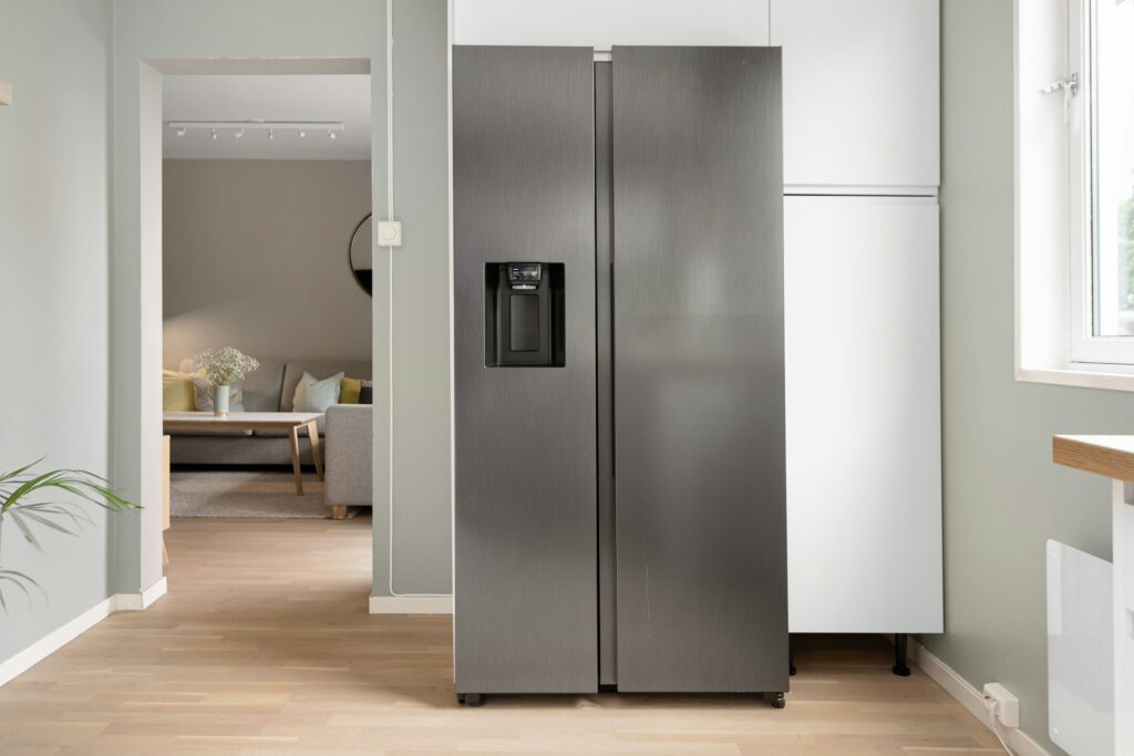 A Modern Stainless Steel Side-By-Side Refrigerator With A Built-In Water And Ice Dispenser, Placed In A Clean, Minimalist Kitchen With Light Wood Flooring And A View Into A Cozy Living Room.