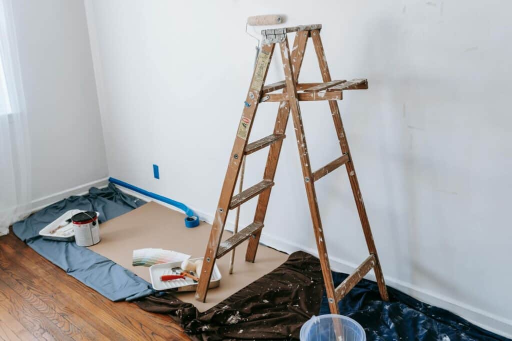 A Wooden Ladder With Paint Splatters Stands In Front Of A White Wall, Surrounded By Painting Supplies Including A Roller, Paint Trays, Blue Painter'S Tape, A Color Swatch Fan, And A Can Of White Paint. The Hardwood Floor Is Protected With Plastic Sheets And Brown Paper.