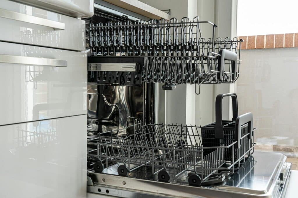 A Close-Up View Of A Modern Stainless Steel Dishwasher With The Door Open, Revealing Two Empty Racks And A Black Utensil Holder. The Surrounding Cabinetry Is White And Glossy, And Natural Light Enters Through A Nearby Window.