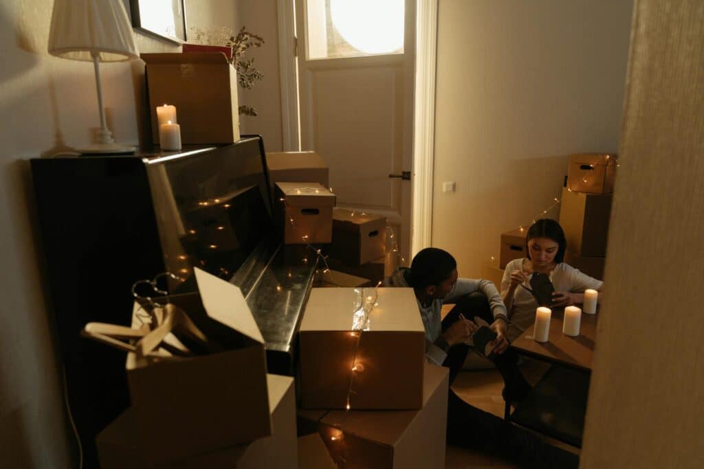 A Cozy Indoor Scene Of A Couple Sitting On The Floor Surrounded By Stacked Moving Boxes In A Dimly Lit Room. Warm Candlelight And String Lights Decorate The Space, With A Piano Against The Wall And A Small Table Holding Candles. The Couple Appears Relaxed While Unpacking.