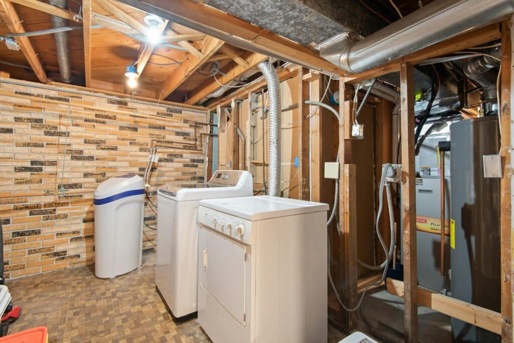 A Detailed Image Of A Basement Laundry And Utility Room With Exposed Wooden Beams, A Washer And Dryer, A Water Softener, Ductwork, And A Water Heater Unit Against A Brick And Wood-Paneled Wall.
