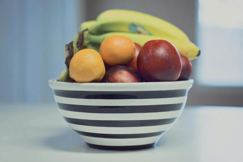 A Black-And-White Striped Ceramic Bowl Filled With Assorted Fresh Fruits, Including Bananas, Red Nectarines, Mandarins, And A Lemon, Sitting On A White Countertop With Soft Natural Light In The Background.