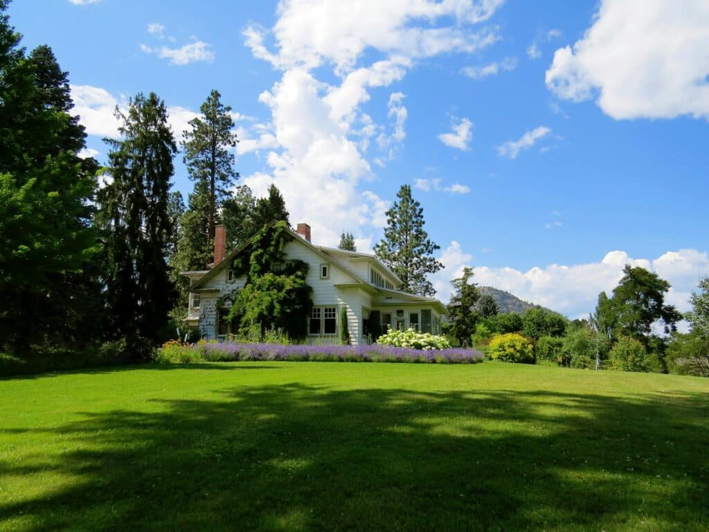 A Scenic Image Of A White House Surrounded By Tall Pine Trees And Colorful Landscaping, With A Large Green Lawn In The Foreground And A Blue Sky Filled With White Clouds In The Background.