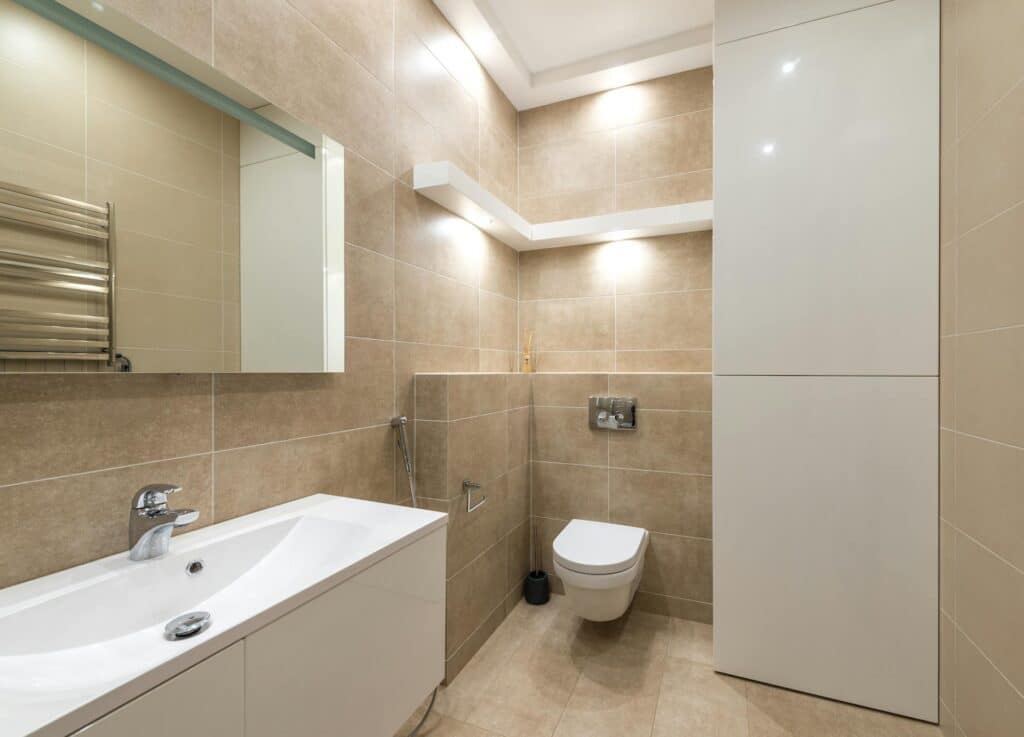 A Modern, Well-Lit Bathroom Featuring Beige Tiled Walls, A Floating White Sink With A Chrome Faucet, A Wall-Mounted Toilet, A Handheld Bidet Sprayer, And Sleek Built-In Cabinetry. Recessed Lighting And A Large Mirror Enhance The Clean, Minimalist Design.