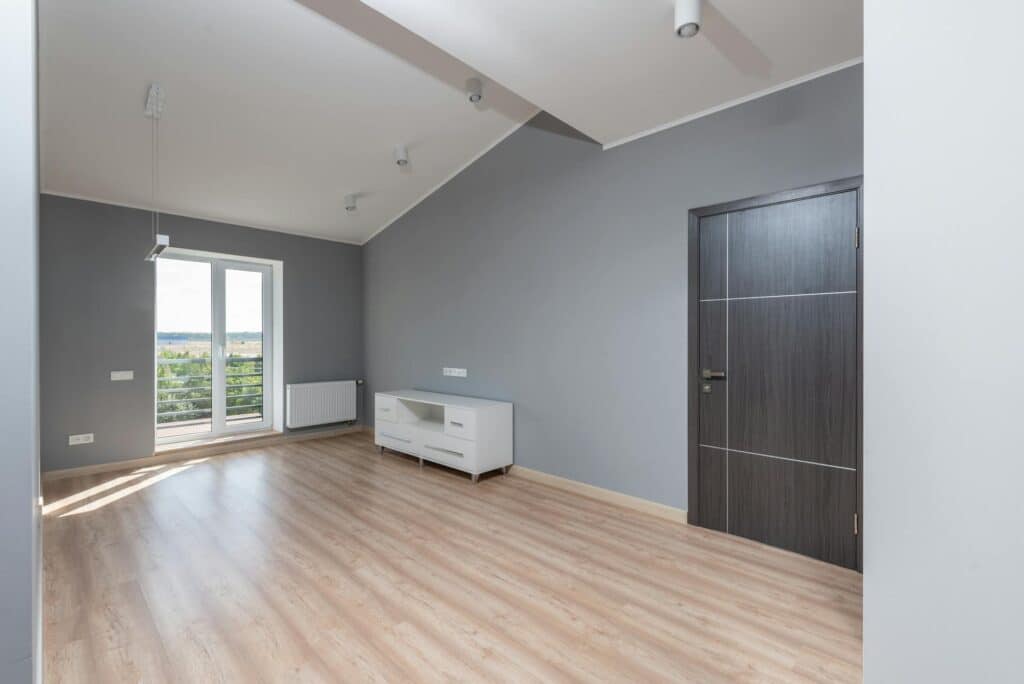 A Modern, Empty Room With Light Wood Flooring, Gray Walls, And A Vaulted White Ceiling With Track Lighting. A Sleek Dark Wood Door Is Closed On The Right Wall, While A White Media Console Sits Below A Row Of Outlets. French Doors Open To A Balcony With A View Of Greenery And A Distant Landscape.