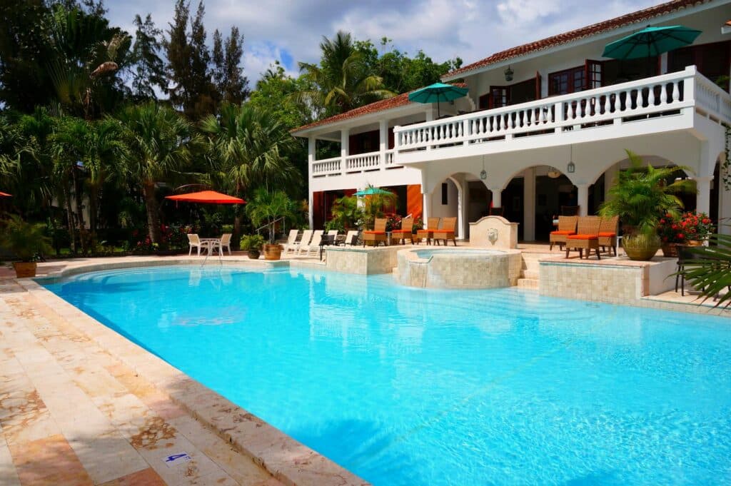 A Large Outdoor Swimming Pool In Front Of A Luxury Villa, Surrounded By Lounge Chairs, Potted Plants, And Umbrellas, With Tropical Trees And Greenery In The Background.