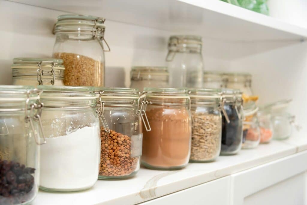 A Neatly Organized Pantry Shelf Lined With Clear Glass Jars Containing Dry Goods Such As Flour, Lentils, Rice, Oats, Cocoa Powder, And Dried Fruit. The Airtight Jars Are Sealed With Metal Clasps To Preserve Freshness And Prevent Pest Intrusion.