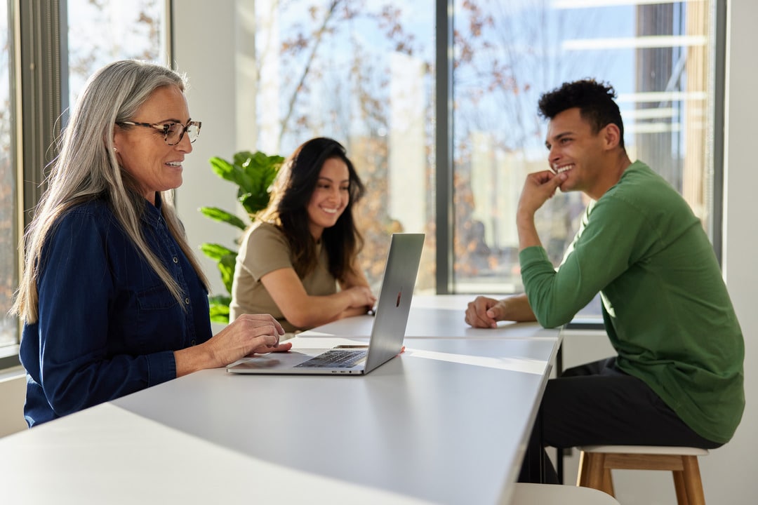 A Diverse Group Of Colleagues At Aptive Pest Control Engaging In A Friendly And Productive Discussion Around A Laptop In A Bright Office Space.