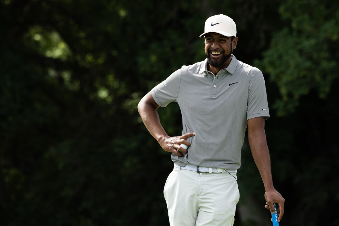 Tony Finau, Wearing A White Cap And A Gray Polo Shirt, Smiles While Holding A Golf Club On A Golf Course, With A Background Of Lush Green Trees.