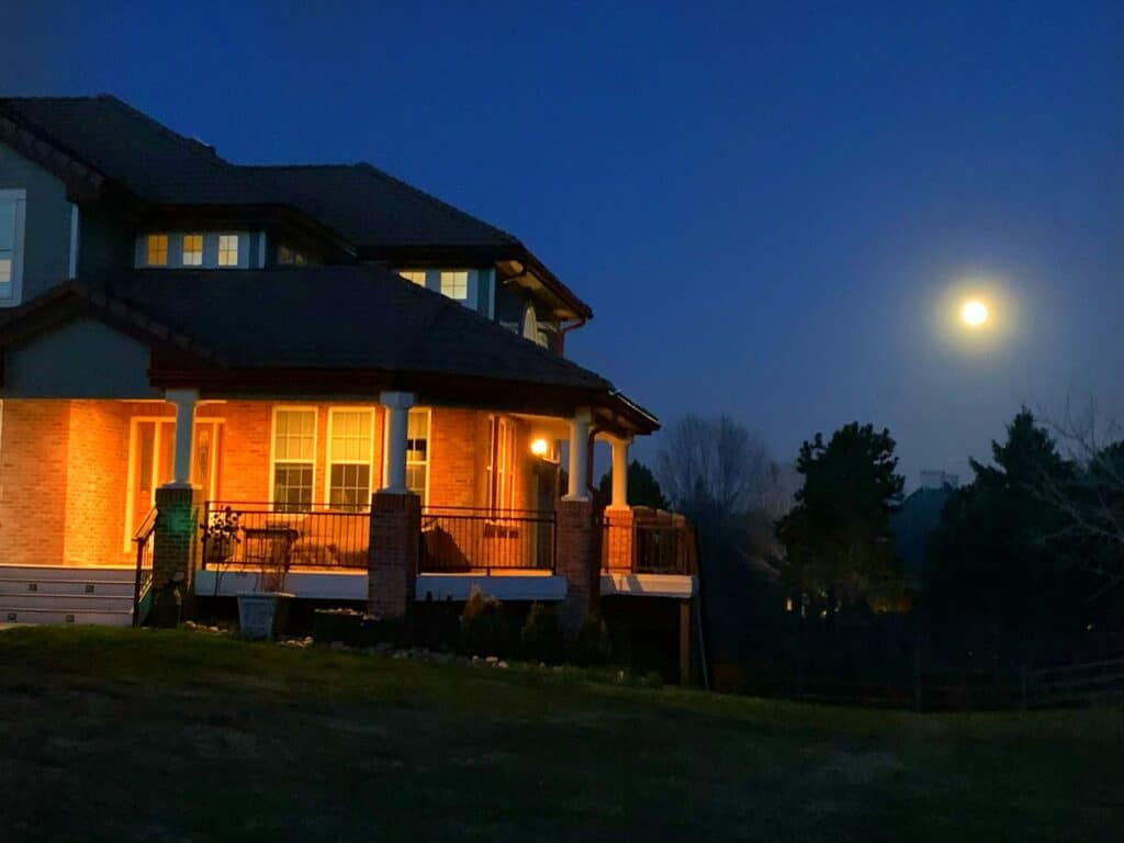 A Warmly Lit House With A Brick Porch And White Pillars At Night, Under A Clear Sky With A Bright Full Moon Rising Above The Tree Line.